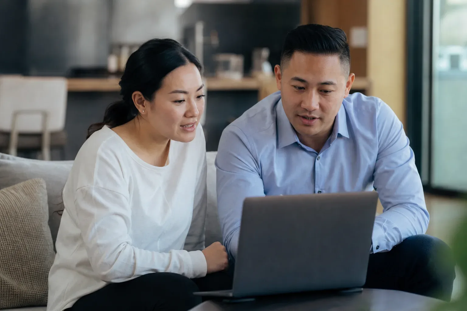 Two people sit together on a sofa, looking at and discussing something on a laptop.