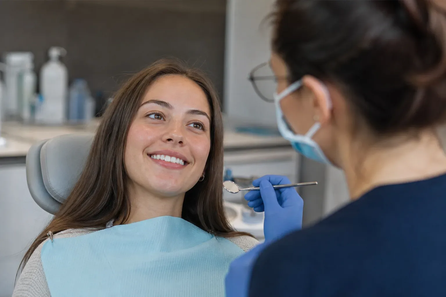 A smiling patient in a dental chair listens to a masked dentist holding a small mouth mirror.