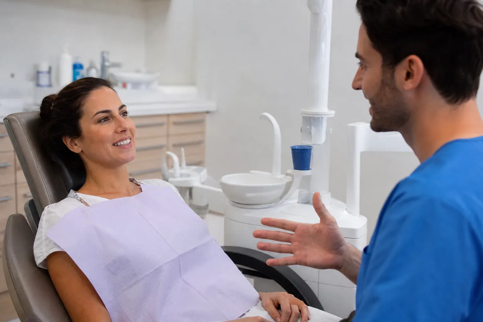 A patient reclines in a dental chair wearing a bib, smiling whilst a dentist in blue scrubs gestures and speaks to them.