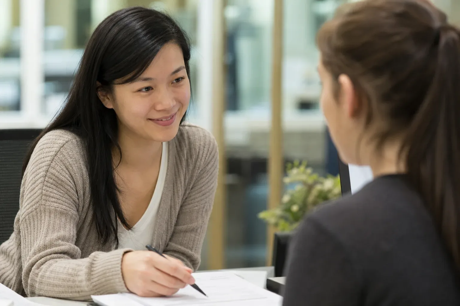 Two people sit across a desk from each other in conversation, one holding a pen over some papers.