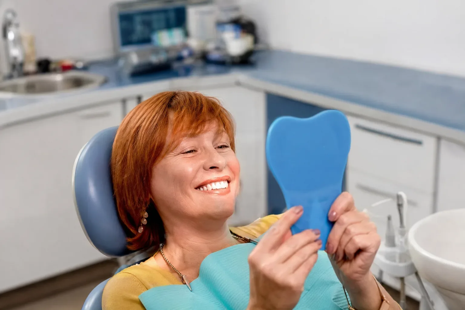 A person sitting in a dental chair smiles whilst looking at their reflection in a tooth-shaped hand mirror.