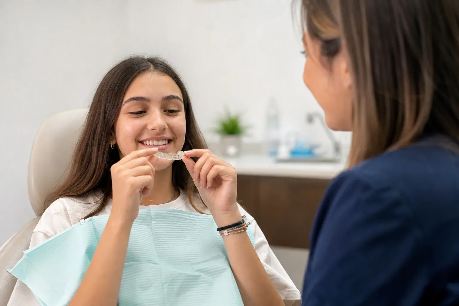 A smiling patient in a dental chair holds a clear dental aligner while a dental professional looks on.