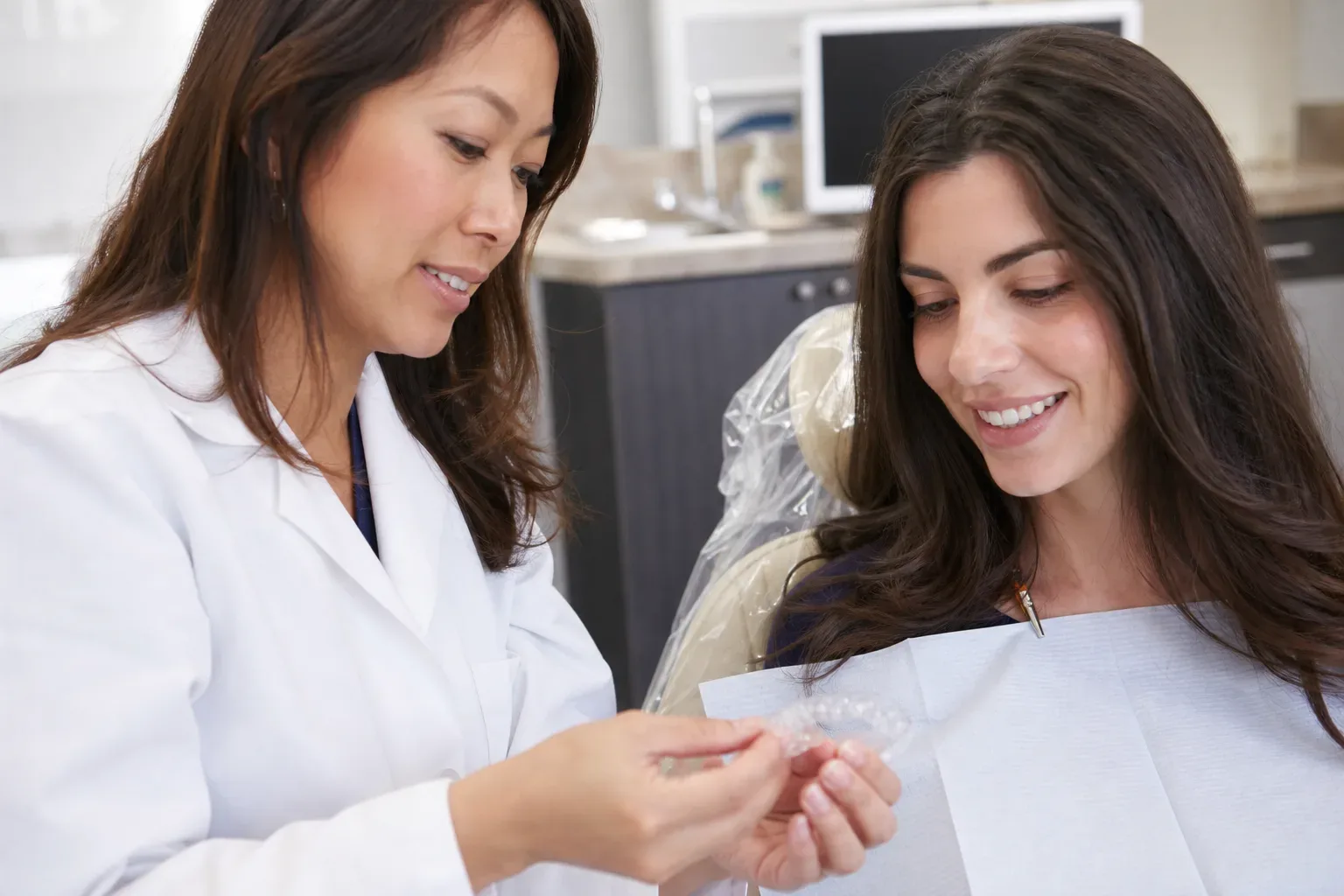 A dentist shows a clear dental aligner to a smiling patient seated in a dental chair.