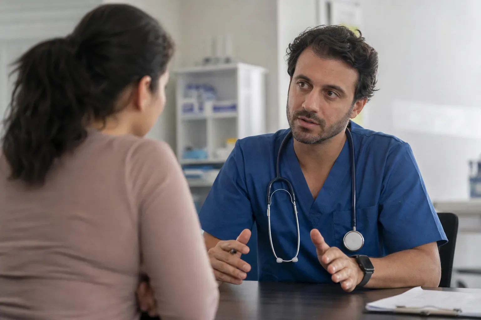 A healthcare worker in blue scrubs and a stethoscope talks with a patient across a desk in a clinical room.