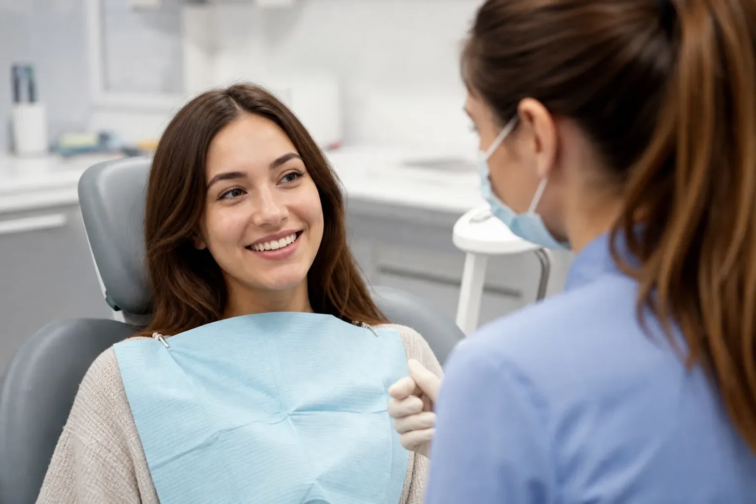 A patient sitting in a dental chair wearing a bib, smiling at a dental professional in scrubs and a face mask.