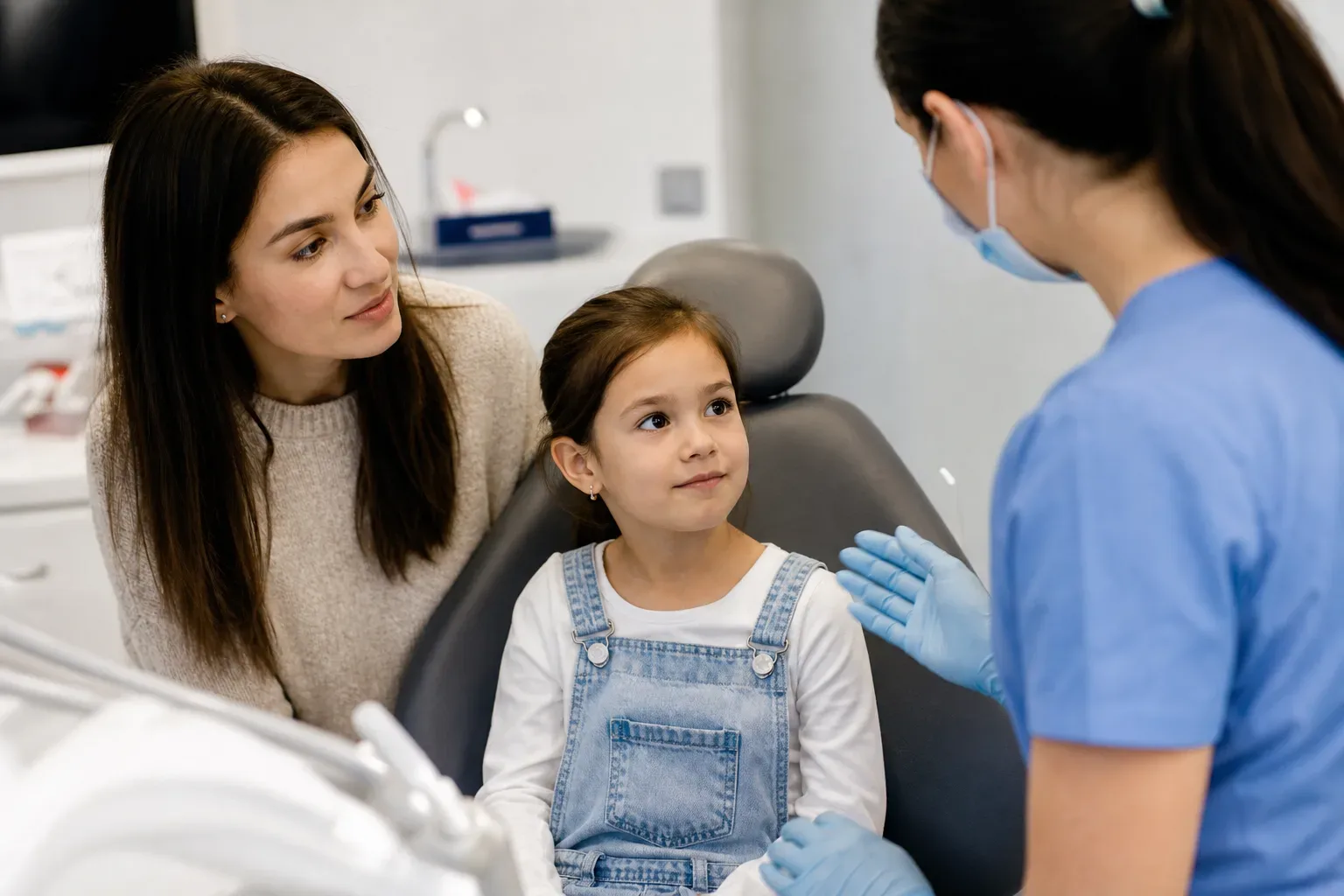 A child sits in a dental chair listening to a dental professional, whilst an adult looks on beside them.