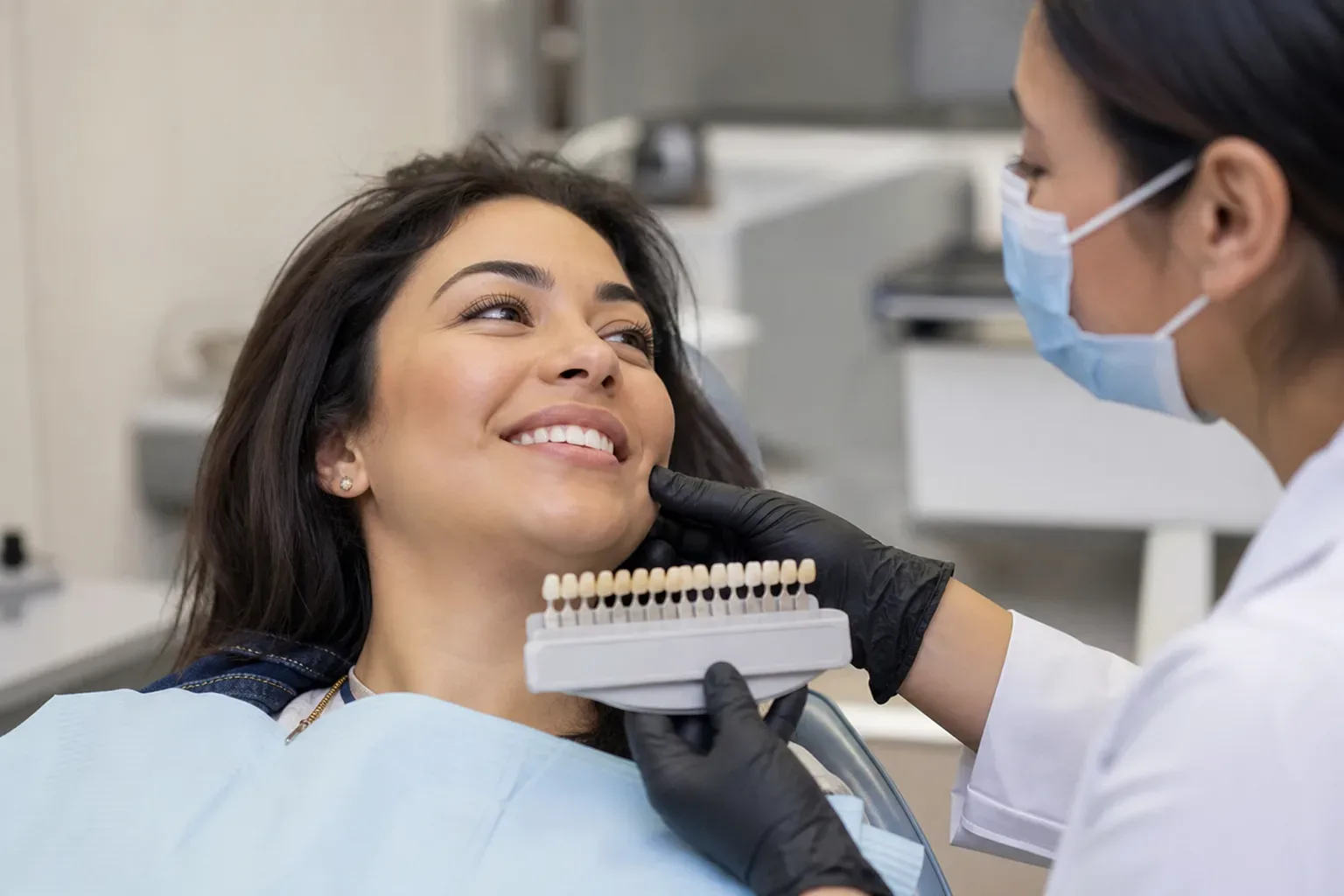 A dental patient smiles as a dentist holds a tooth shade guide up to their face in a clinic.