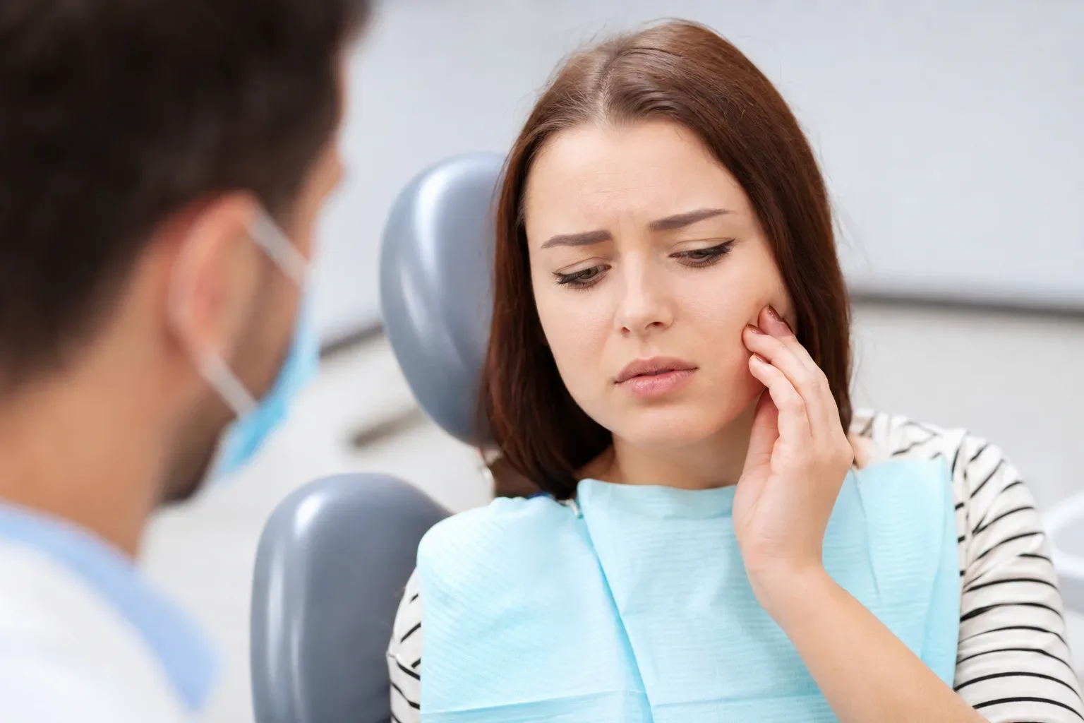 A patient holding their cheek in pain sits in a dental chair while a masked dentist listens.