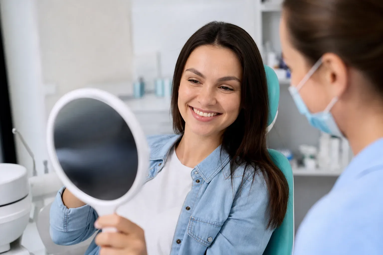 A patient sits in a dental chair holding a mirror and smiling, while a masked dental professional looks on.