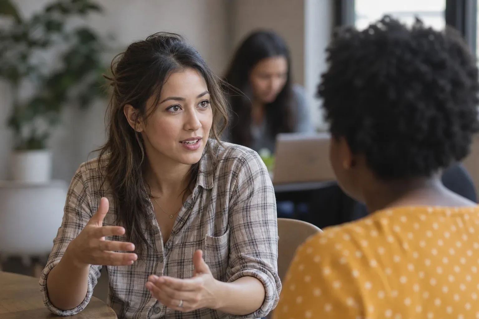 Two people sit talking at a table in a busy open workspace, one gesturing expressively with both hands.
